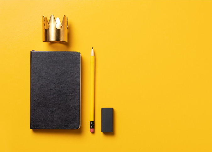 a black notebook laying on a bright yellow backdrop, with a crown above the notebook, and a pencil laying to the right
