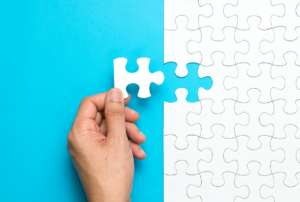 image of a hand holding a white puzzle piece on a bright blue background, poised to add the final piece to a larger, complete puzzle