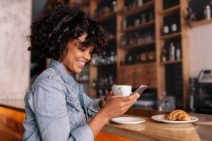 pretty african american woman sitting a coffee bar holding her smartphone and a cup of coffee